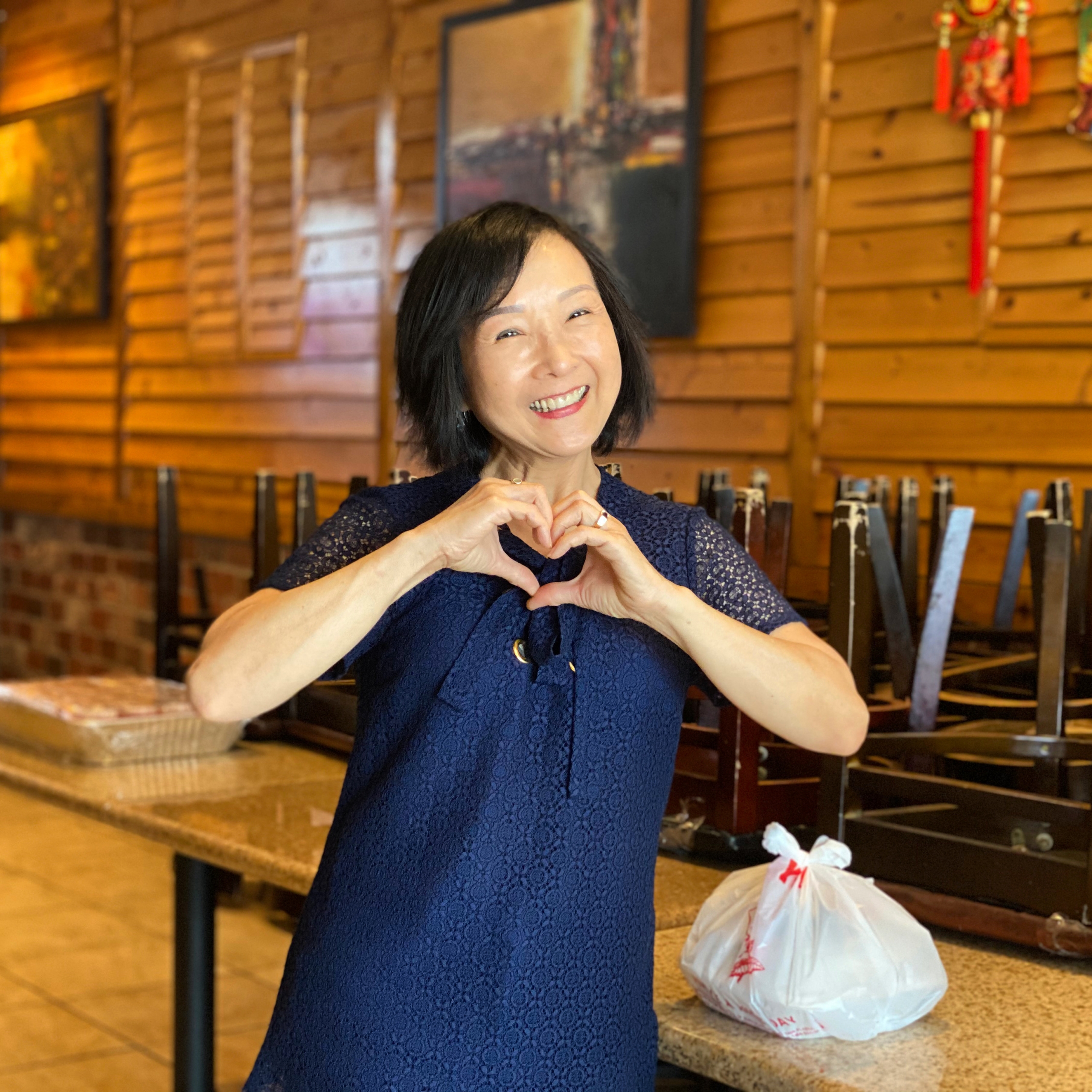 Co-owner Trinh Ong makes a heart shape with her hands, standing next to a takeout order in her restaurant.
