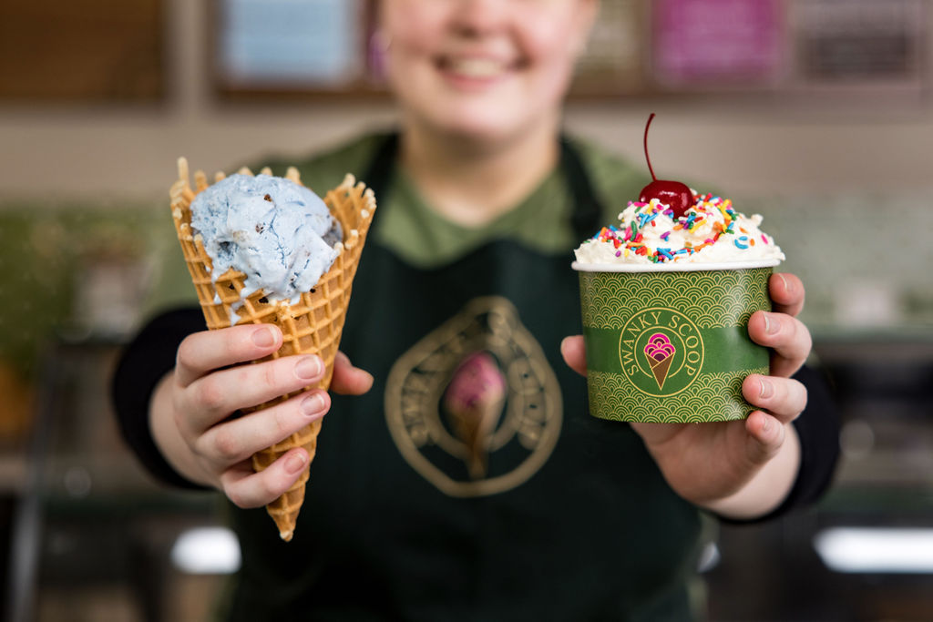 An ice cream cone and a cup of ice cream being held by an employee at Swanky Scoop