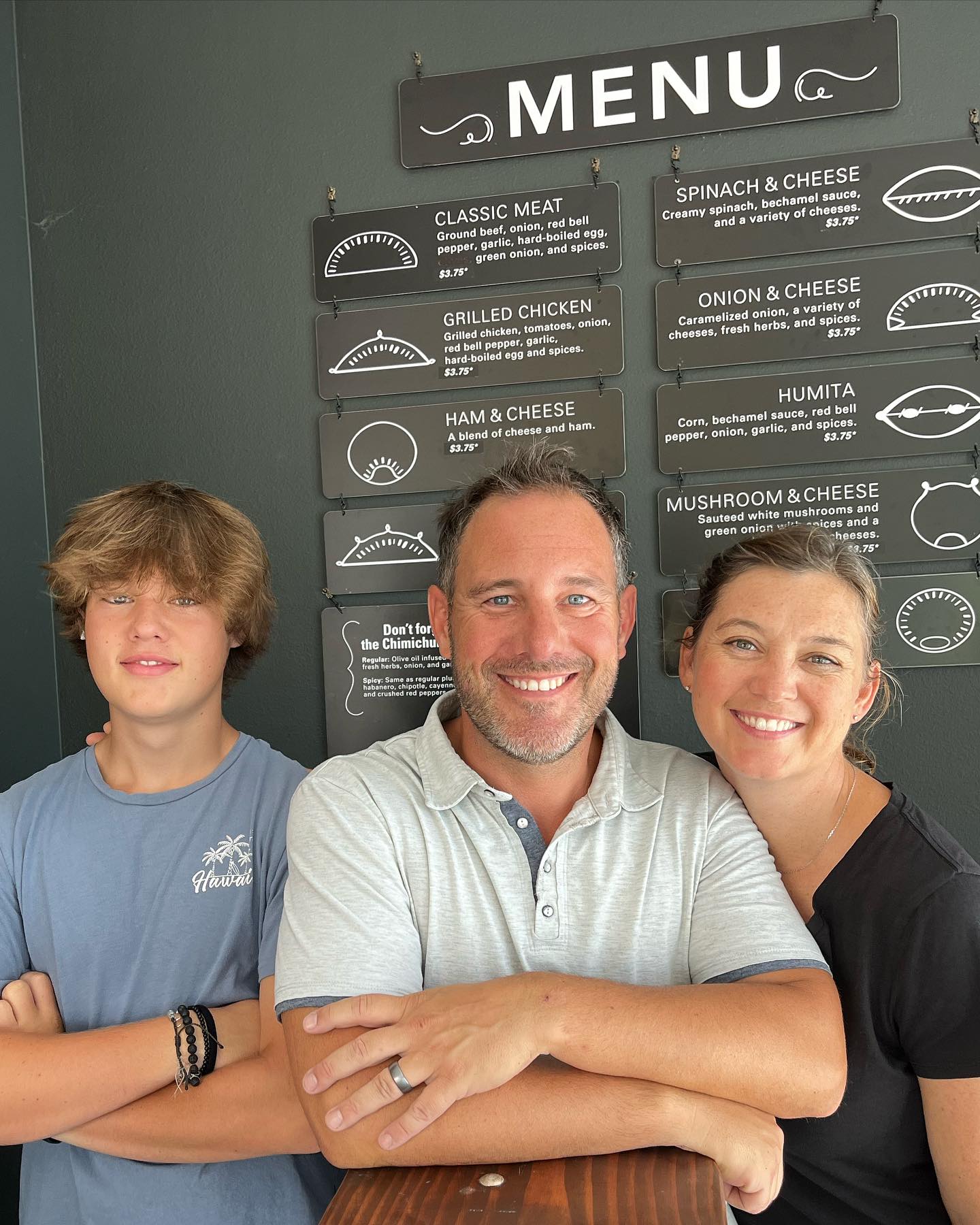 Rocky's Emanadas family owners husband, wife, and son, standing in front of the empanada menu