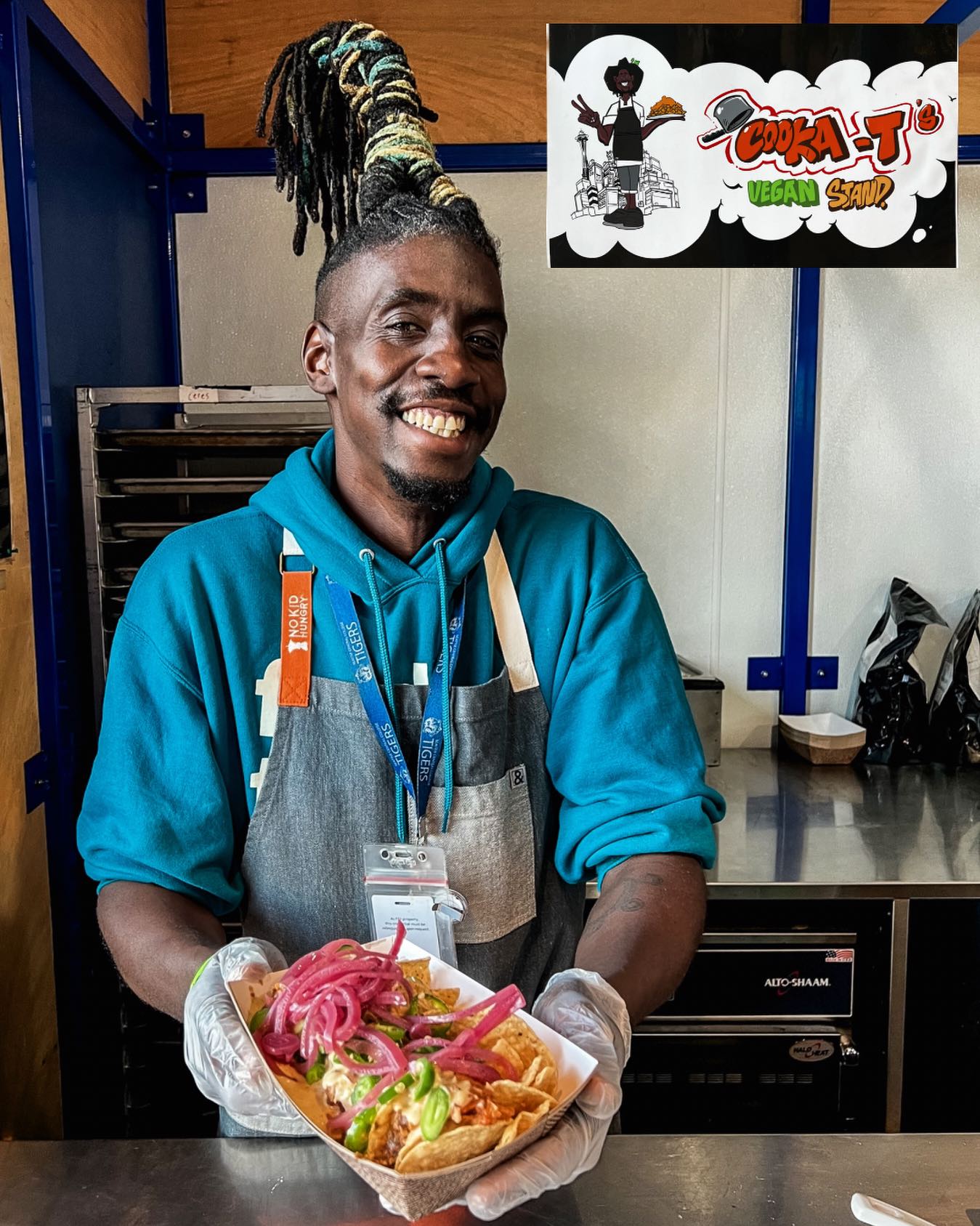 Cooka T standing in front of his food stall at Lumen Field holding a plate of his vegan nachos